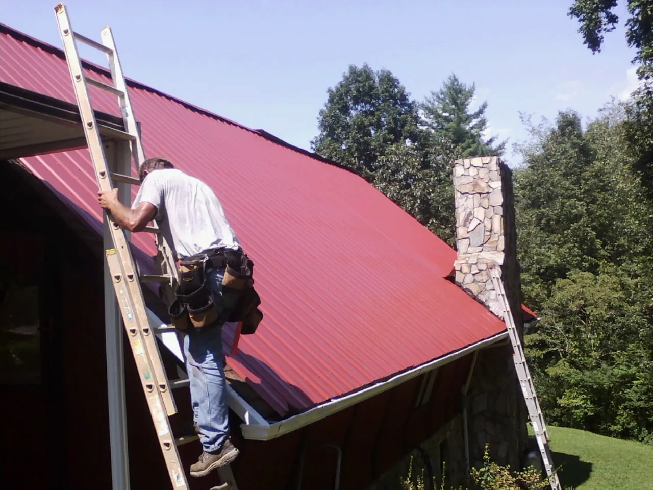 Roofer on a ladder beside a red metal roof and stone chimney — Soffit & Fascia Repair by Roofing Brothers in Mount Croghan